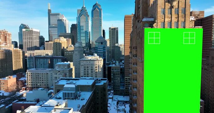 A tall green screen wrap covers the flank of a brick building above snowy rooftops in Philadelphia, Pennsylvania, the Center City towers rising behind it. Aerial lateral wide shot