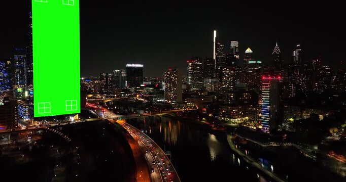A tall green screen wrap glows on a building beside the Schuylkill at night in Philadelphia, Pennsylvania, expressway light trails and the lit Center City skyline beyond. Aerial view