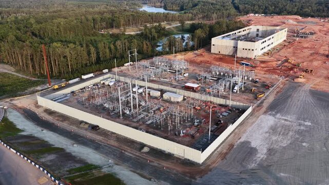Walled substation and its control building sit beside a data center campus in Louisa County, Virginia, with graded clay, a woodland pond and pine forest running to the horizon. Aerial rising shot
