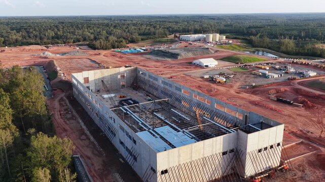 A yellow crawler crane sets roof steel inside a data hall shell in Louisa County, Virginia, ringed by red clay pads, white work tents and storage tanks against deep pine forest. Aerial wide shot