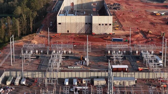 Rows of steel switchgear and a blank concrete electrical building stand in a new substation in Louisa County, Virginia, with excavators and trailers working the red clay apron. Aerial view