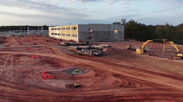 Braced end wall of a data hall under construction in Louisa County, Virginia, with an excavator, loaded flatbed trailers and a wheel loader working the red clay around it. Aerial approaching shot