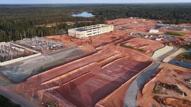 Terraced red clay pads and haul roads wait for the next data halls in Louisa County, Virginia, with the substation and a long two story building standing at the treeline. Aerial wide shot