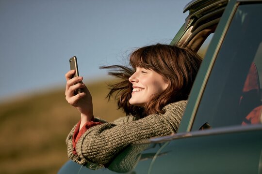 Happy young woman leaning out of a car window holding a smartphone during a road trip adventure