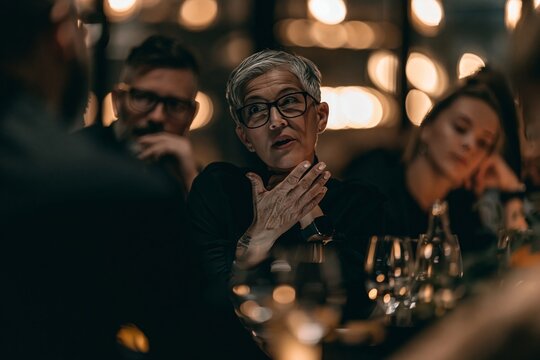 Mature woman with short grey hair and glasses speaking passionately during a business dinner