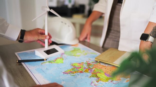 Two renewable energy engineers discussing a global project in their office, using a world map and a wind turbine model to plan the international expansion of their wind farm
