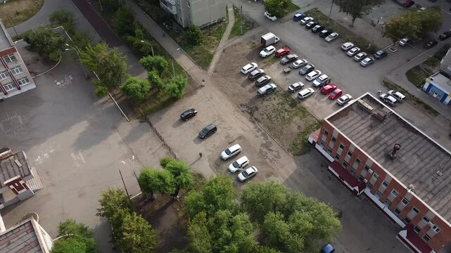 Aerial view of a parking lot near apartment buildings. Cars parked in rows, some scattered. Trees and patches of grass separate the spaces. Distant buildings form a residential backdrop