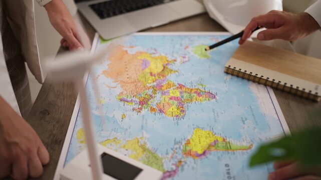 Engineers planning wind turbine installations using a world map to identify suitable regions for renewable power generation, discussing wind farm development and sustainable electricity production.