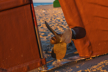 Bronze boat propeller framed between rudder and hull on sandy sunset beach © Roman Bjuty