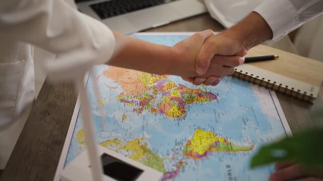 Two renewable energy engineers finalizing an international green power deal, shaking hands over a world map with a miniature wind turbine model spinning in the foreground on the office desk