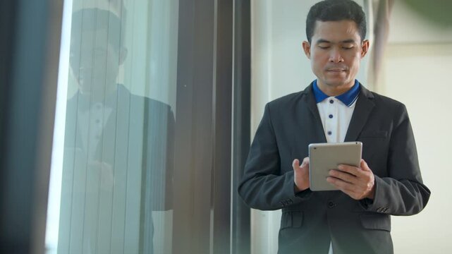 Professional man in a suit jacket using a digital tablet while standing near a glass window