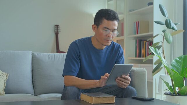 Focused man in glasses using a digital tablet while sitting on a comfortable living room sofa