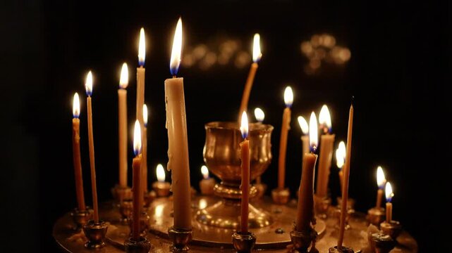 Many thin beeswax candles burning on a circular metal stand inside a dark church interior. Slow motion