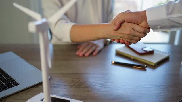 Close-up view of a businessman and a businesswoman shaking hands in an office, finalizing an agreement on a wind energy project with a model wind turbine on the desk