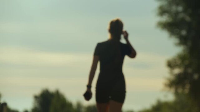 Woman walking away along park path with raised hand under sky, soft green trees in calm light sunset