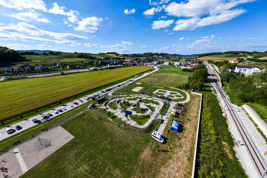 A wide aerial view of the asphalt pumptrack park in Pesnica pri Mariboru, Slovenia, surrounded by green fields, rolling hills, a railway line, and a bright blue sky filled with fluffy white clouds.