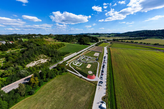 A wide aerial view of the asphalt pumptrack park in Pesnica pri Mariboru, Slovenia, surrounded by green fields, rolling hills, a railway line, and a bright blue sky filled with fluffy white clouds.