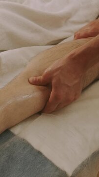 close-up hands massage calf with oil sheen, thumbs pressing during manual therapy warm light on skin and towel