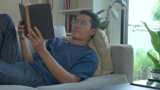 A focused man wearing glasses relaxes on a comfortable sofa while reading a hardcover book
