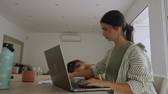 Working mother typing on laptop with daughter playing nearby