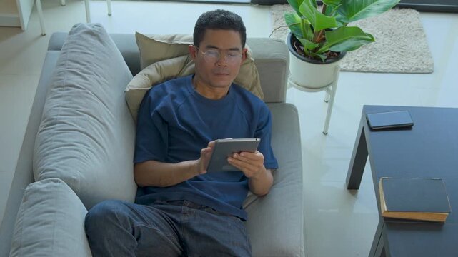 Asian man relaxing on a sofa while using a digital tablet in a bright modern living room space