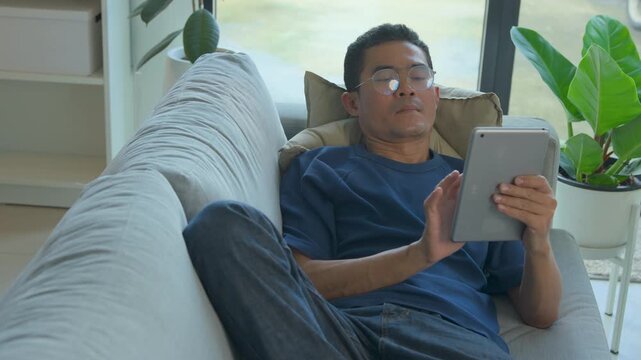 Asian man relaxing on a sofa while using a digital tablet in a bright modern living room space