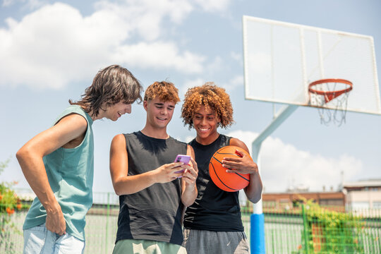 Diverse teen friends sharing smartphone on basketball court