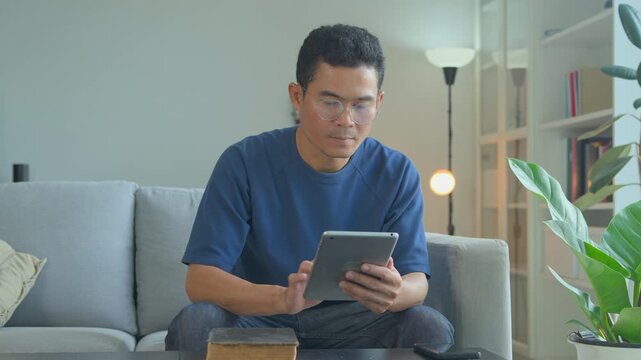 Focused man in glasses using a digital tablet while sitting on a comfortable living room sofa