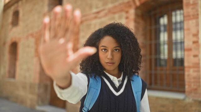 Woman student in school uniform and backpack raises hand palm forward in a stopping gesture against a brick building facade; determination.