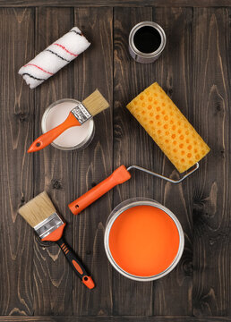 High-angle studio shot of open paint buckets with white, orange, and black colors. Professional painter tools, paintbrushes, and rollers arranged on a dark wood workshop table.