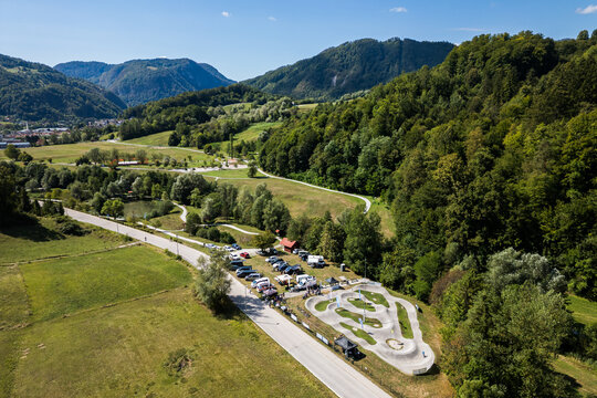 A wide aerial view of the asphalt pumptrack park in Zagorje ob Savi, Slovenia, surrounded by green fields, rolling hills, a railway line, and a bright blue sky filled with fluffy white clouds.