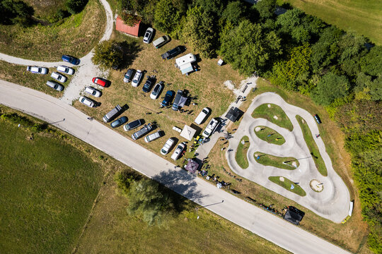 A wide aerial view of the asphalt pumptrack park in Zagorje ob Savi, Slovenia, surrounded by green fields, rolling hills, a railway line, and a bright blue sky filled with fluffy white clouds.