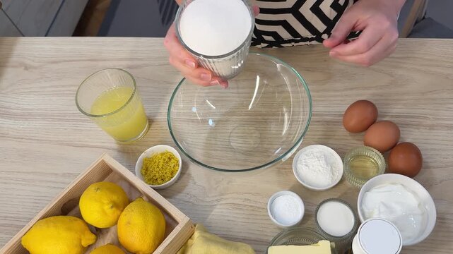 A person pours white granulated sugar from a glass cup into a large clear glass mixing bowl on a light wooden table surrounded by lemons, eggs, and flour