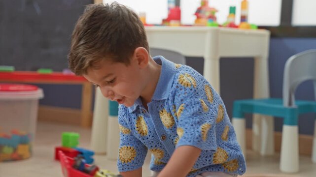 Boy in blue crocheted sweater pushing a toy car and reaching for stacked blocks while kneeling on playroom floor in building; quiet curiosity.