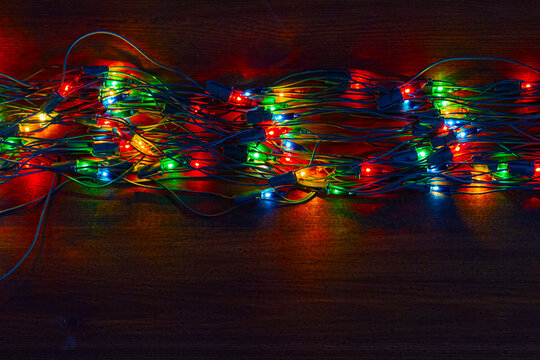 A colorful fairy lights garland lies across a dark wooden surface. Multiple glowing bulbs create intricate tangles of wire. Holiday celebration and festive seasonal decor for interior party