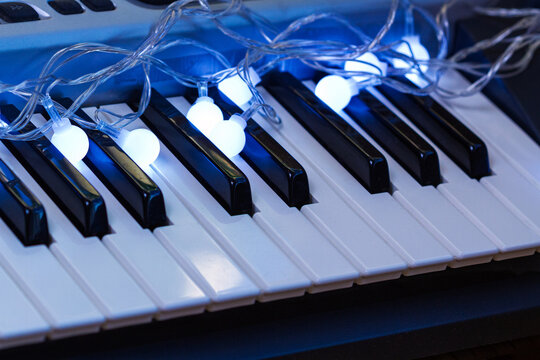 A white led bulb garland rests across the black and white keys of a music keyboard. The small round bulbs emit a bright blue glow over the instrument. Christmas musical celebration