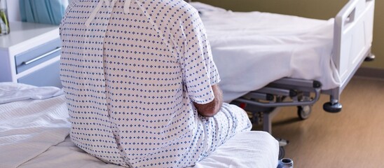 A patient wearing a hospital gown sits alone on the edge of a hospital bed, seen from behind....