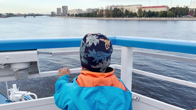 A young boy looks out curiously from a cruise ship railing over the Neva River. He feels excited and full of wonder. The peaceful river view brings a joyful travel moment.