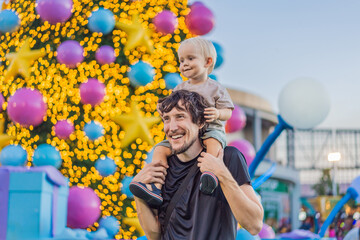 Fototapeta premium Dad smiles with a three year old child among colorful holiday decorations, showing fatherhood, family celebration and festive outing concept