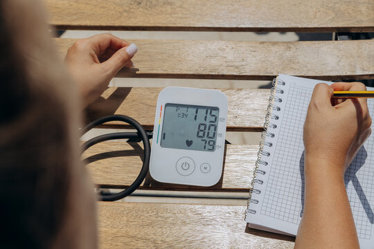 Blood Pressure Monitor On Table With Person Recording Readings In Notebook For Home Health Tracking