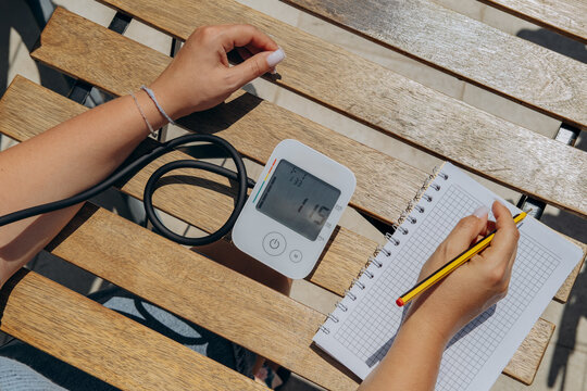 Woman Recording Blood Pressure Reading With Digital Monitor On Wooden Table Outdoors