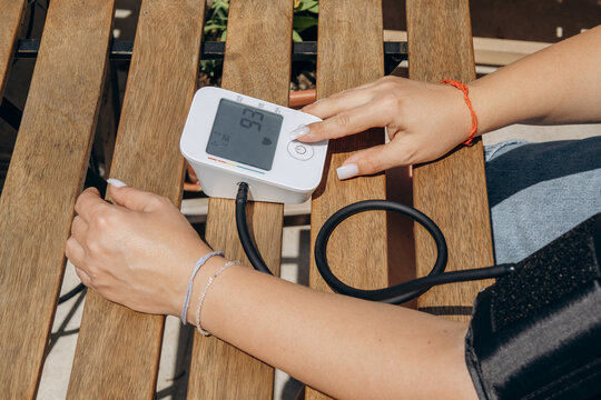 Young Woman Checking Blood Pressure With Digital Monitor On Outdoor Wooden Bench