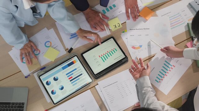 Overhead view of multiple hands pointing at financial charts on a tablet, surrounded by printed reports and colorful data visualizations on a busy desk.