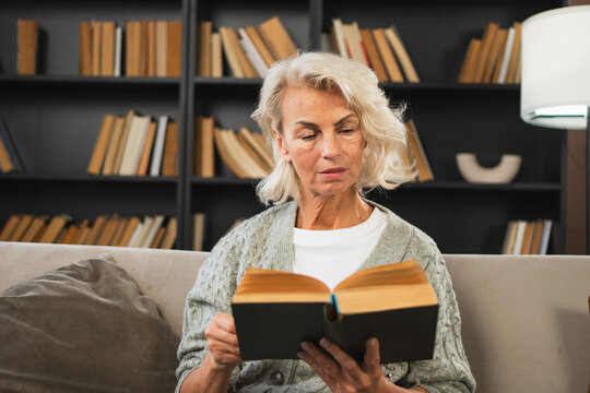 Comfort relaxed senior old elderly woman reading book at home. Mature woman resting enjoy calm day sitting on sofa at home alone. Adult lady spending free time keeping book smiling. Pension concept