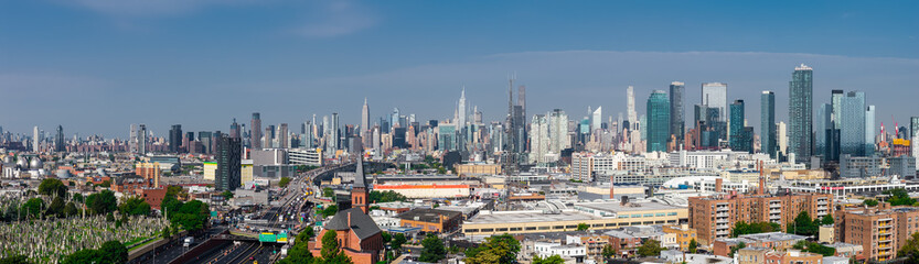 Obraz premium Aerial wide panorama captures the distant Manhattan skyscrapers rising beyond Queens neighborhoods, cemetery grounds, and highway traffic on a clear day.