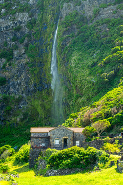A stunning view of the 90-meter high Cascata do Poco do Bacalhau waterfall cascading down a lush green volcanic cliff right behind a traditional rustic stone house in Faja Grande, Flores Island