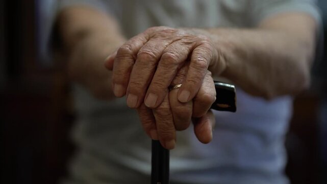 Elderly 80-year-old man gently rests both wrinkled hands on a dark cane, capturing age, strength and quiet dignity.