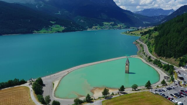 Aerial drone forward flight over the turquoise Lake Reschen, approaching the submerged church bell tower and coastal road in South Tyrol, Italy
