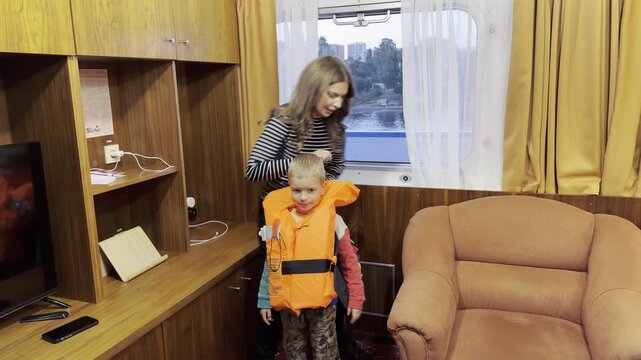 A mother helps her happy son put on an orange lifejacket in a cruise ship cabin on the Neva River, Saint Petersburg. The boy laughs with joyful excitement. They feel safe and cozy.