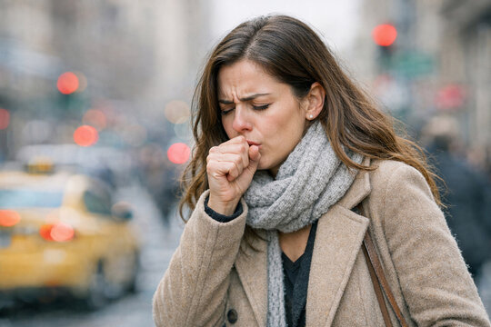 A woman in a cityscape coughs and sneezes due to air pollution and allergens.